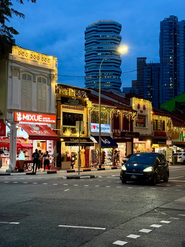 A Singapore street at dusk, shophouses lit up beneath high-rise towers